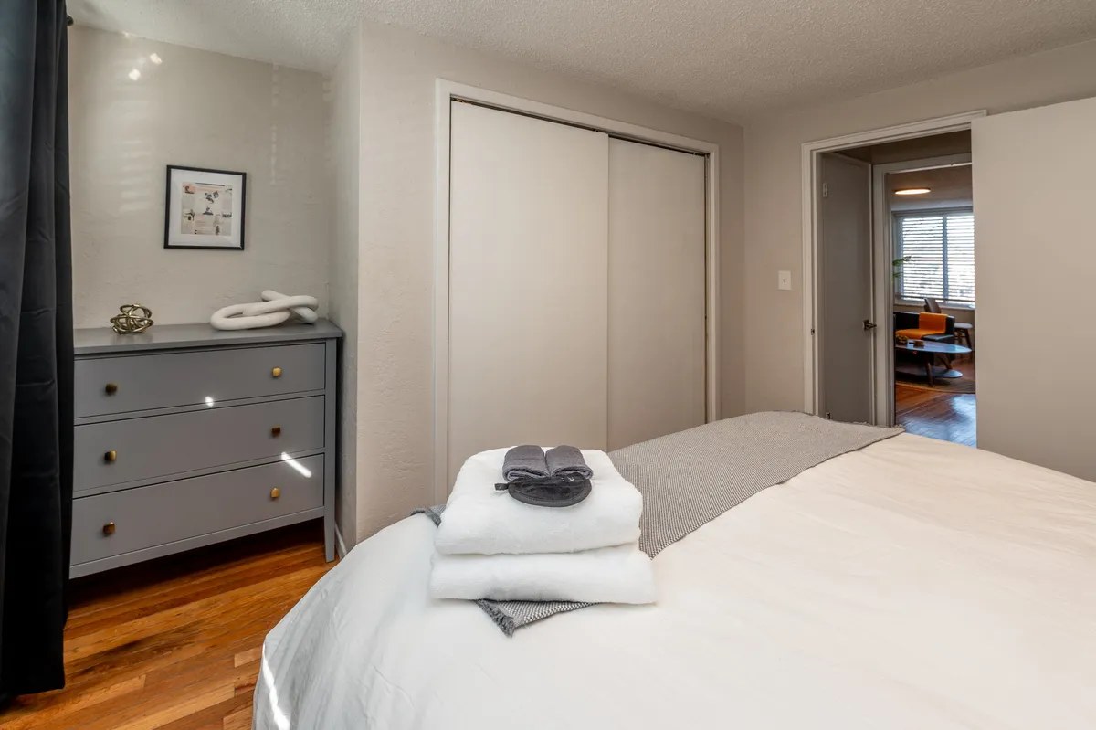 A neatly arranged bed with crisp white linens anchors this peaceful bedroom. A chest of drawers provides storage, while subtle decorative wall art creates a serene and harmonious environment. Sunlight softly brightens the space, reinforcing the room's calm ambiance.