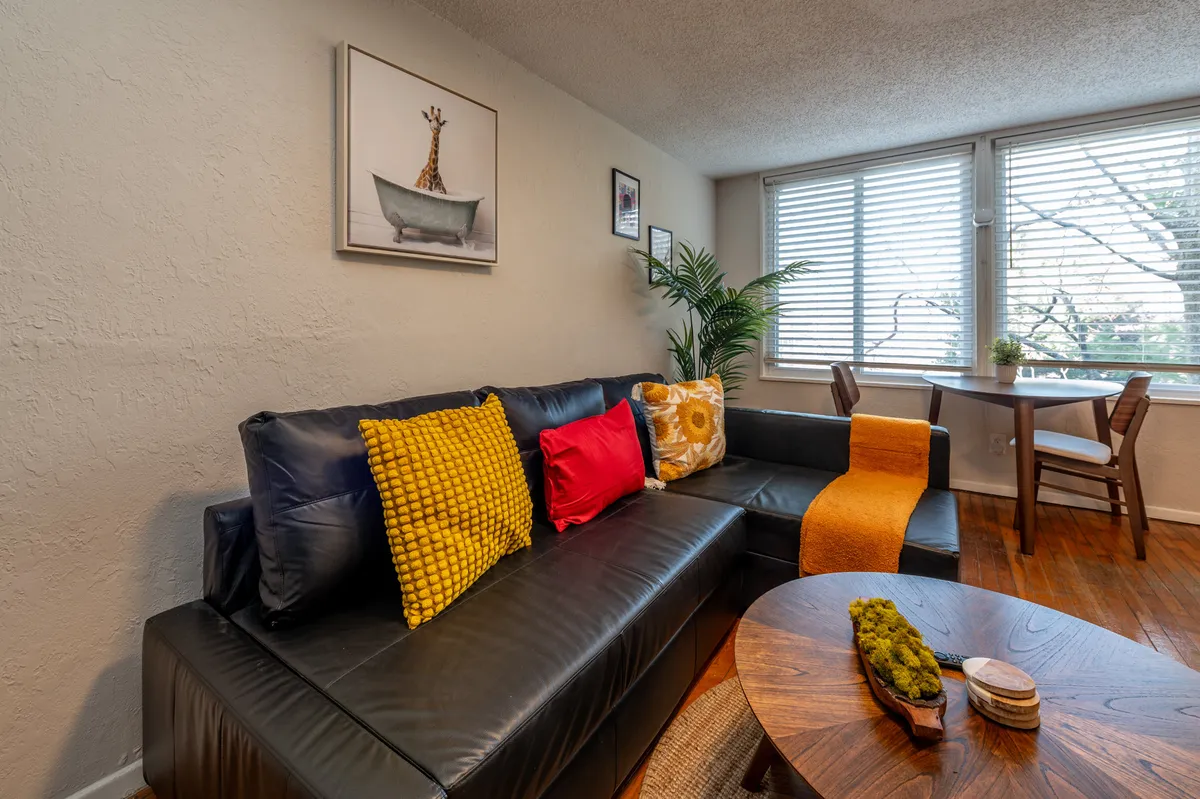 A cozy living area featuring a plush sofa with soft cushions, a stylish coffee table, decorative artwork on the walls, and a large window that fills the space with natural light. The arrangement encourages both relaxation and social interaction, with plenty of daylight streaming in.