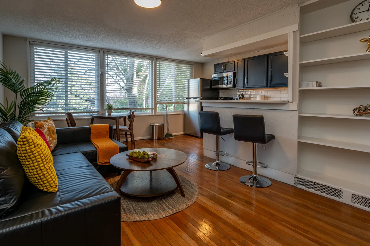 An inviting open-plan space that seamlessly blends the living area with a kitchen and dining table. The sofa with cushions faces a round coffee table, while the kitchen and dining area are visible in the background, suggesting versatility and adaptability for different daily routines.