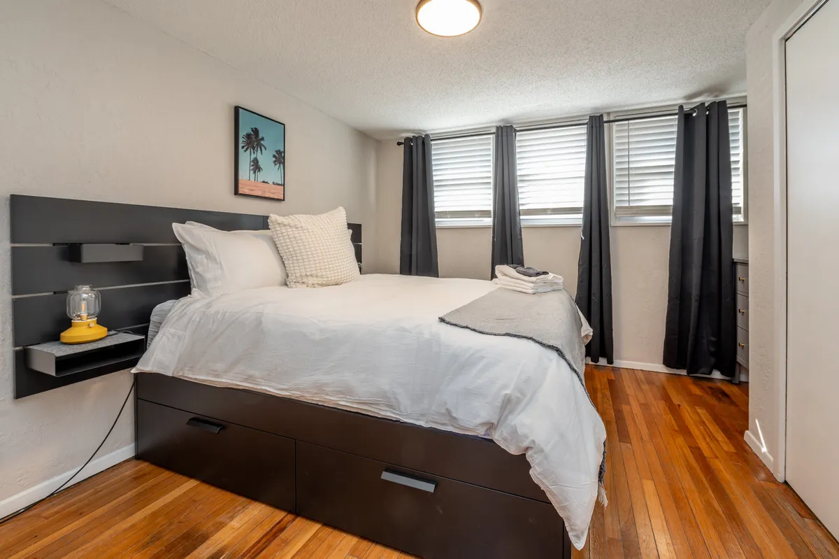 This image features a cozy bedroom setting with a plush bed, a nightstand, and window curtains. Natural light gently filters into the room, while a framed photo adds a personal touch. The layout suggests a relaxing atmosphere perfect for reading or unwinding.